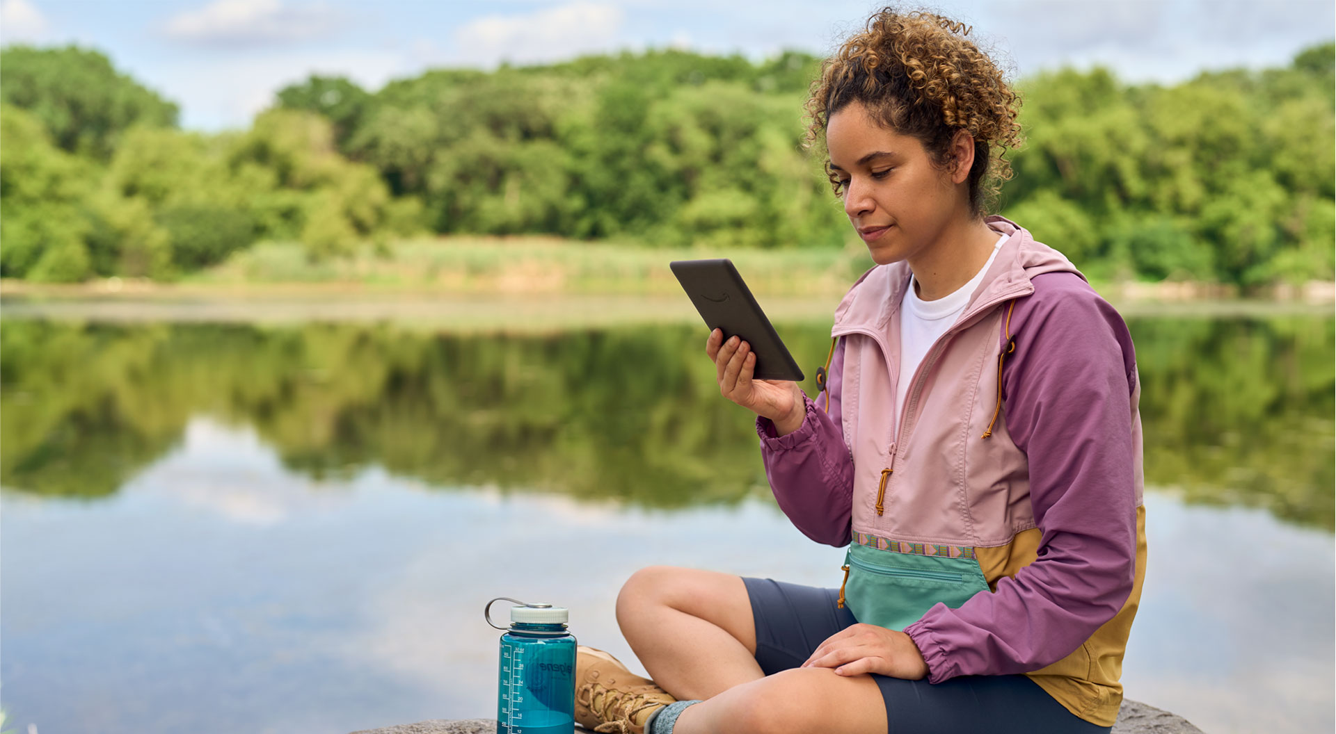 Woman reads a Kindle Paperwhite while sitting by a lake.